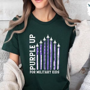 Op de afbeelding: Een donkergroen T-shirt met een witte afbeelding van "Purple Up" voor militaire kinderen. De afbeelding toont een rij vliegtuigen met paarse strepen en witte sterren.