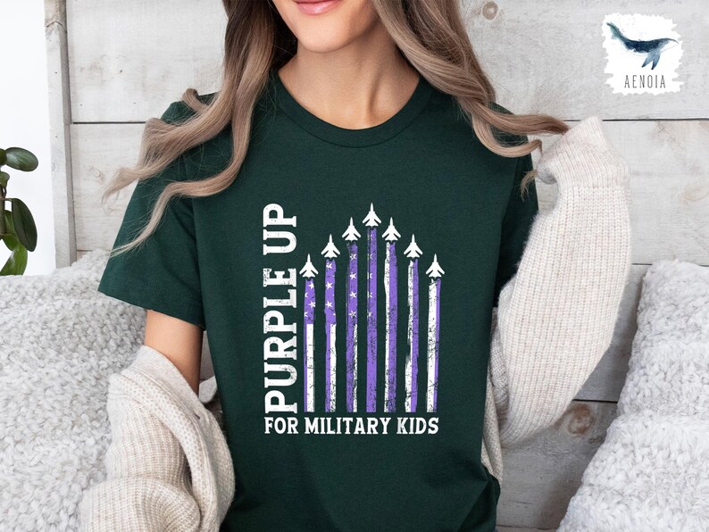 Op de afbeelding: Een donkergroen T-shirt met een witte afbeelding van "Purple Up" voor militaire kinderen. De afbeelding toont een rij vliegtuigen met paarse strepen en witte sterren.