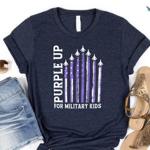 Op de afbeelding: Een marineblauw T-shirt met een witte afbeelding van een paarse Amerikaanse vlag met vliegtuigen die eroverheen vliegen. De tekst "Purple Up for Military Kids" staat onder de vlag gedrukt.