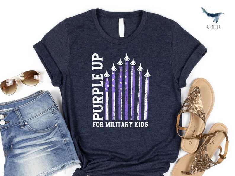 Op de afbeelding: Een marineblauw T-shirt met een witte afbeelding van een paarse Amerikaanse vlag met vliegtuigen die eroverheen vliegen. De tekst "Purple Up for Military Kids" staat onder de vlag gedrukt.