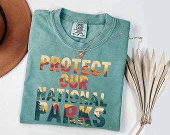 May include: A teal Comfort Colors t-shirt with the words "PROTECT OUR NATIONAL PARKS" in a colorful, layered design. A silver necklace with a pendant and a dried plant are next to the shirt. The shirt is small, size S-P-CH.