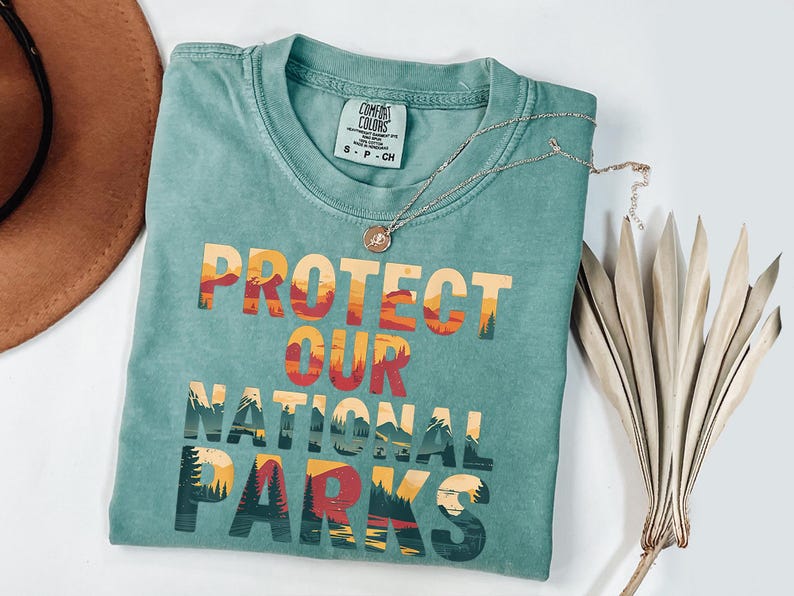May include: A teal Comfort Colors t-shirt with the words "PROTECT OUR NATIONAL PARKS" in a colourful, layered design. A silver necklace with a pendant and a dried plant are next to the shirt. The shirt is small, size S-P-CH.