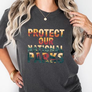May include: Dark grey t-shirt featuring the text "PROTECT OUR NATIONAL PARKS" in a colourful, nature-themed design. The shirt has a crew neck and short sleeves. The design includes a mountain and forest landscape.