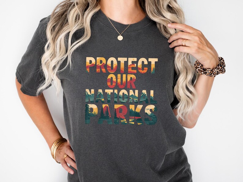 May include: Dark grey t-shirt featuring the text "PROTECT OUR NATIONAL PARKS" in a colourful, nature-themed design. The shirt has a crew neck and short sleeves. The design includes a mountain and forest landscape.