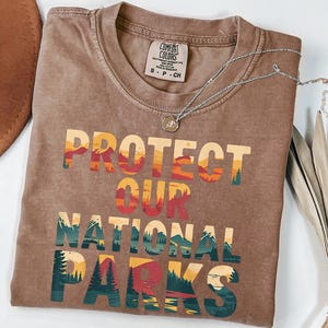 May include: A brown t-shirt featuring the text "PROTECT OUR NATIONAL PARKS" in a layered, colourful design with mountain and forest illustrations. A silver necklace and a brown hat are also visible.