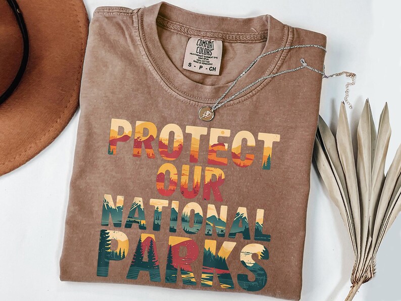 May include: A brown t-shirt featuring the text "PROTECT OUR NATIONAL PARKS" in a layered, colourful design with mountain and forest illustrations. A silver necklace and a brown hat are also visible.