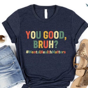 以下が含まれることがあります： カラフルな文字で「YOU GOOD, BRUH?」とハッシュタグ#MentalHealthMattersがプリントされたネイビーブルーのTシャツ。デニムショーツ、サングラス、サンダルと組み合わせています。