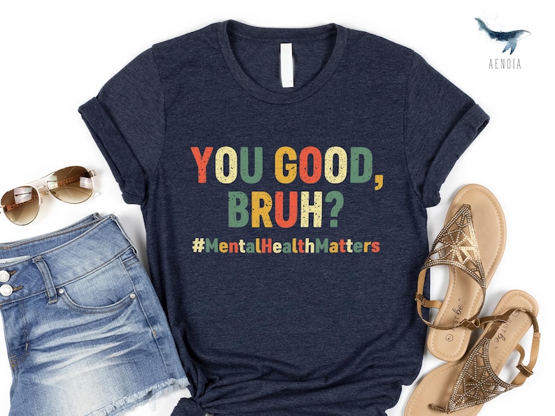 以下が含まれることがあります： カラフルな文字で「YOU GOOD, BRUH?」とハッシュタグ#MentalHealthMattersがプリントされたネイビーブルーのTシャツ。デニムショーツ、サングラス、サンダルと組み合わせています。