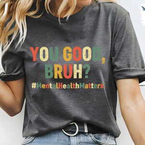 以下が含まれることがあります： 「YOU GOOD, BRUH?」とカラフルな文字で書かれた濃いグレーのTシャツ。テキストの下にはハッシュタグ「#MentalHealthMatters」があります。このシャツはカジュアルなスタイルです。