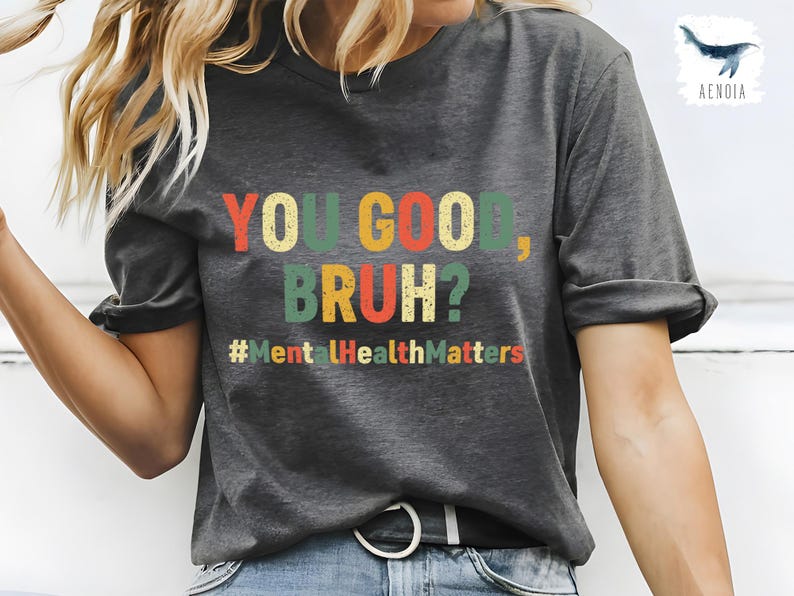 以下が含まれることがあります： 「YOU GOOD, BRUH?」とカラフルな文字で書かれた濃いグレーのTシャツ。テキストの下にはハッシュタグ「#MentalHealthMatters」があります。このシャツはカジュアルなスタイルです。
