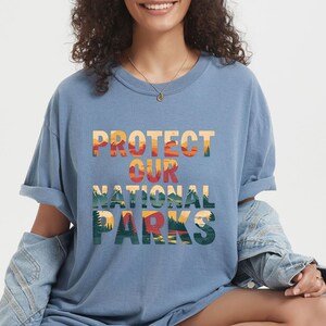 May include: A blue t-shirt with the words "PROTECT OUR NATIONAL PARKS" in a colourful, layered design. The text features a landscape illustration with trees, mountains, and a sunset. The shirt has rolled-up sleeves.