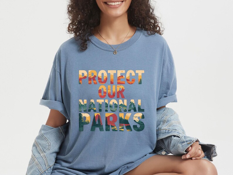 May include: A blue t-shirt with the words "PROTECT OUR NATIONAL PARKS" in a colourful, layered design. The text features a landscape illustration with trees, mountains, and a sunset. The shirt has rolled-up sleeves.