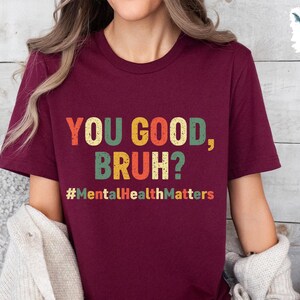 以下が含まれることがあります： レトロなスタイルのカラフルな文字で「YOU GOOD, BRUH?」と書かれたバーガンディ色のTシャツ。その下にハッシュタグ「#MentalHealthMatters」があります。このシャツはクルーネックです。