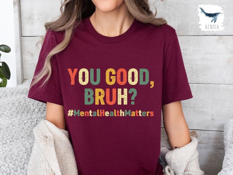 以下が含まれることがあります： レトロなスタイルのカラフルな文字で「YOU GOOD, BRUH?」と書かれたバーガンディ色のTシャツ。その下にハッシュタグ「#MentalHealthMatters」があります。このシャツはクルーネックです。