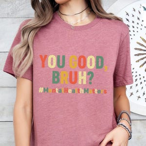以下が含まれることがあります： レトロな色使いの「YOU GOOD, BRUH?」の文字がプリントされた、くすんだピンク色のTシャツ。テキストの下には「#MentalHealthMatters」のハッシュタグ。半袖のクルーネックTシャツです。