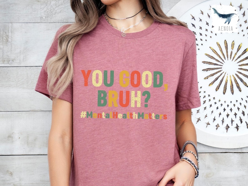以下が含まれることがあります： レトロな色使いの「YOU GOOD, BRUH?」の文字がプリントされた、くすんだピンク色のTシャツ。テキストの下には「#MentalHealthMatters」のハッシュタグ。半袖のクルーネックTシャツです。