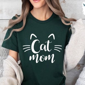 May include: A dark green t-shirt with the words "Cat Mom" in white script, featuring cat ears and whiskers. The shirt is a crew neck style. A cream-coloured cardigan is draped over the shoulders.