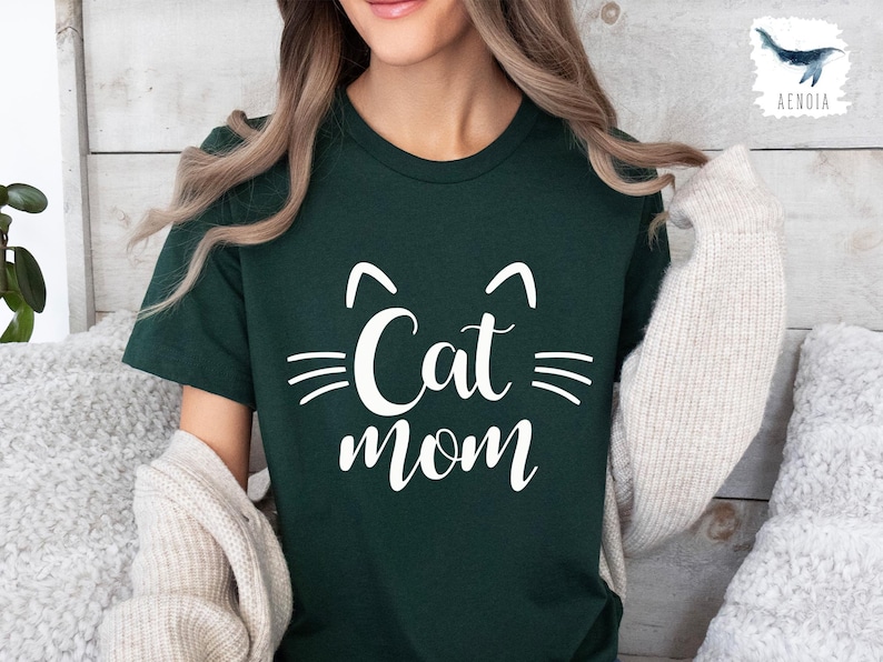 May include: A dark green t-shirt with the words "Cat Mom" in white script, featuring cat ears and whiskers. The shirt is a crew neck style. A cream-coloured cardigan is draped over the shoulders.