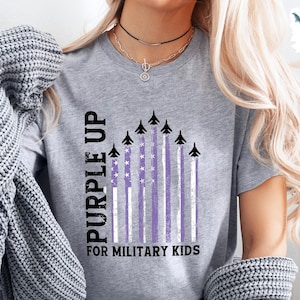 Op de afbeelding: Een grijs T-shirt met de tekst "Purple Up for Military Kids" en een afbeelding van paarse en witte strepen met vliegtuigen die eroverheen vliegen.