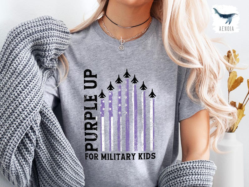 Op de afbeelding: Een grijs T-shirt met de tekst "Purple Up for Military Kids" en een afbeelding van paarse en witte strepen met vliegtuigen die eroverheen vliegen.