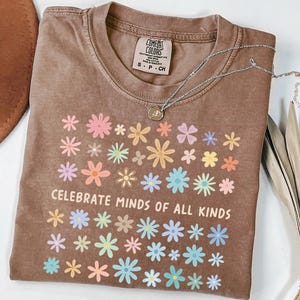 May include: A brown t-shirt with a floral design and the text "CELEBRATE MINDS OF ALL KINDS". The shirt has a crew neck and short sleeves. A silver necklace with a pendant is on top of the shirt.