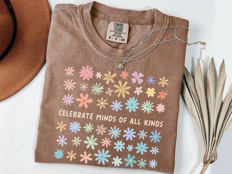 May include: A brown t-shirt with a floral design and the text "CELEBRATE MINDS OF ALL KINDS". The shirt has a crew neck and short sleeves. A silver necklace with a pendant is on top of the shirt.