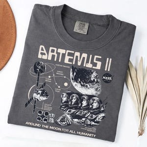 May include: Dark gray t-shirt featuring the text "ARTEMIS II" with a graphic of astronauts, a moon, and a spacecraft. The NASA logo is also visible. The phrase "AROUND THE MOON FOR ALL HUMANITY" is printed at the bottom.