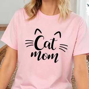 May include: A pale pink t-shirt featuring a black cat face design and the words "Cat Mom" in a script font. The shirt has short sleeves and a crew neck. The design includes cat ears and whiskers.