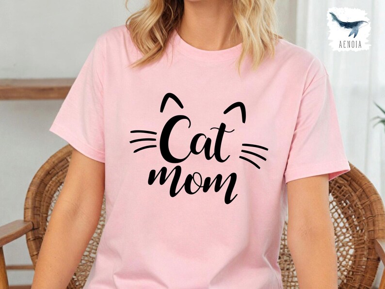 May include: A pale pink t-shirt featuring a black cat face design and the words "Cat Mom" in a script font. The shirt has short sleeves and a crew neck. The design includes cat ears and whiskers.