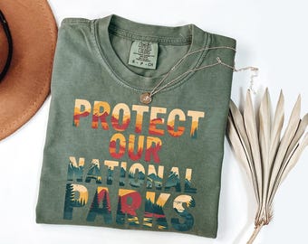 May include: Olive green t-shirt with the text "PROTECT OUR NATIONAL PARKS" in a colorful, layered design. A silver necklace with a pendant rests on the shirt. A brown hat and dried plant are also in the image.