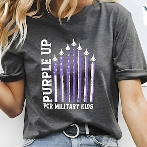 Op de afbeelding: Een donkergrijs T-shirt met een 'Purple Up for Military Kids'-afbeelding. De afbeelding toont een Amerikaanse vlag in paars en wit met vliegtuigen die eroverheen vliegen.