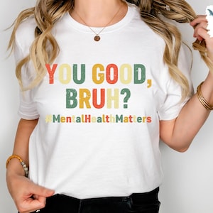 以下が含まれることがあります： レトロなフォントで「YOU GOOD, BRUH？」と書かれた白いTシャツ。シャツには、カラフルなヴィンテージスタイルのハッシュタグ「#MentalHealthMatters」も含まれています。カジュアルな普段着のTシャツ。