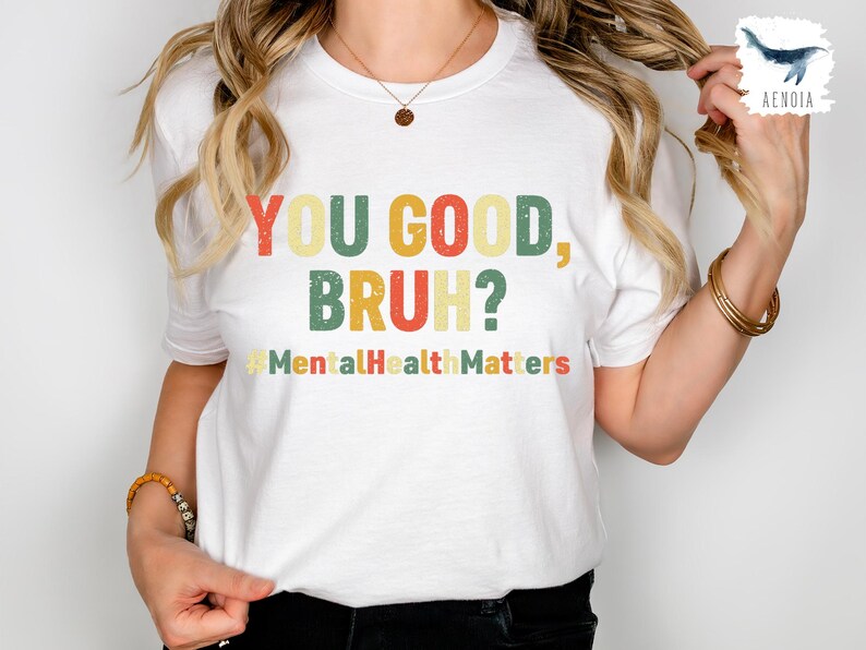 以下が含まれることがあります： レトロなフォントで「YOU GOOD, BRUH？」と書かれた白いTシャツ。シャツには、カラフルなヴィンテージスタイルのハッシュタグ「#MentalHealthMatters」も含まれています。カジュアルな普段着のTシャツ。