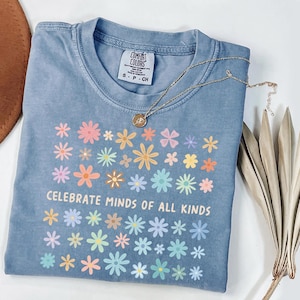 May include: A light blue t-shirt with a floral design in pastel colors and the text "CELEBRATE MINDS OF ALL KINDS". A gold necklace with a pendant is on the shirt. A brown hat and dried plant are in the background.