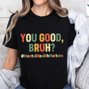 以下が含まれることがあります： レトロカラーで「YOU GOOD, BRUH？」と書かれた黒いTシャツ。その下に「#MentalHealthMatters」のハッシュタグがあります。普段使いにぴったりの、快適でカジュアルなトップスです。