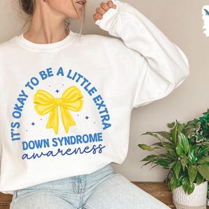 May include: White sweatshirt with a blue and yellow graphic that says "It's okay to be a little extra Down Syndrome awareness".