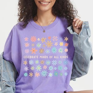 May include: A lilac-colored t-shirt with a floral design and the text "Celebrate Minds of All Kinds." The shirt features a crew neck and short sleeves. The model is wearing a denim jacket.