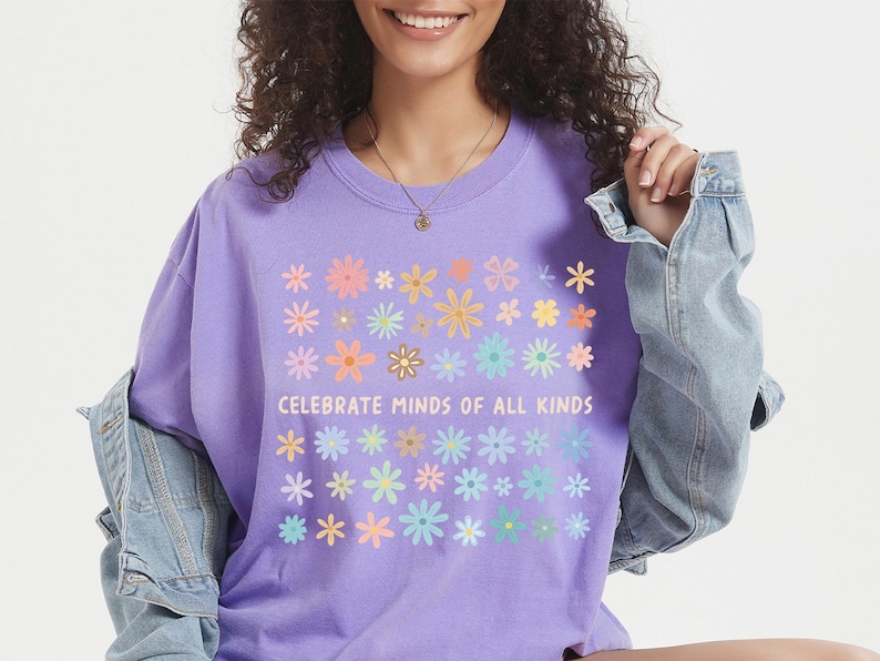 May include: A lilac-colored t-shirt with a floral design and the text "Celebrate Minds of All Kinds." The shirt features a crew neck and short sleeves. The model is wearing a denim jacket.