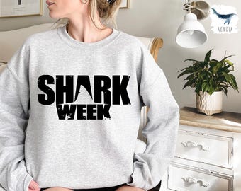 May include: A light gray sweatshirt with the words "SHARK WEEK" printed in large, black, distressed font. The design features a shark fin incorporated into the letter "A". The sweatshirt is a casual, comfortable style.