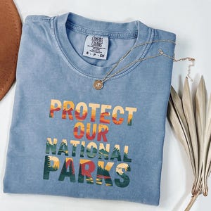 May include: A blue Comfort Colors t-shirt with the text "PROTECT OUR NATIONAL PARKS" in a colourful, layered design. A gold necklace with a pendant rests on the shirt. A brown hat and dried plant are in the background.