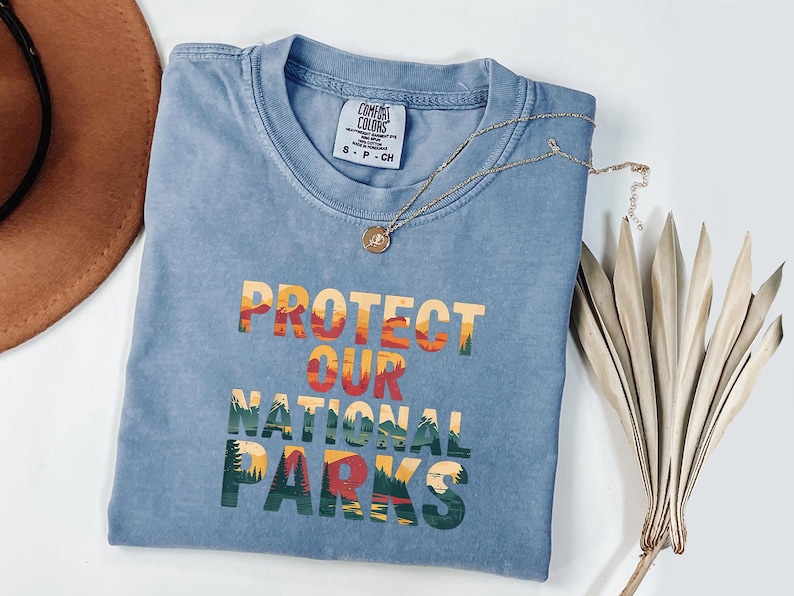 May include: A blue Comfort Colors t-shirt with the text "PROTECT OUR NATIONAL PARKS" in a colourful, layered design. A gold necklace with a pendant rests on the shirt. A brown hat and dried plant are in the background.