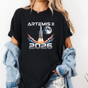 May include: Black t-shirt featuring a graphic of a rocket launching into space, with the text "ARTEMIS II 2026". The design includes a moon, stars, and colorful vapor trails. The shirt is a casual wear item.