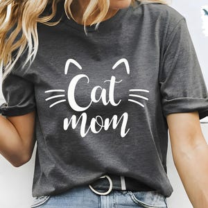 May include: Dark grey t-shirt featuring the text "Cat mom" in white, with cat ear and whisker graphics. The shirt is a casual style, suitable for everyday wear. The design is simple and bold.