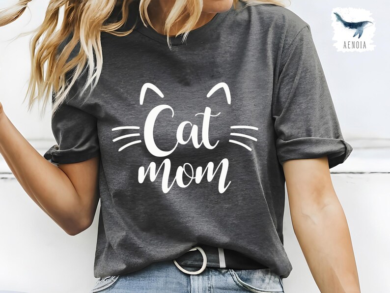 May include: Dark grey t-shirt featuring the text "Cat mom" in white, with cat ear and whisker graphics. The shirt is a casual style, suitable for everyday wear. The design is simple and bold.