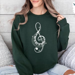 Peut inclure: Un sweat-shirt vert fonc&eacute; avec un motif de cl&eacute; de sol blanche. La cl&eacute; de sol est compos&eacute;e de touches de piano et de volutes.