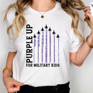 Op de afbeelding: Wit T-shirt met een paarse opdruk voor militaire kinderen. De opdruk toont een paarse Amerikaanse vlag met vliegtuigen die eroverheen vliegen.