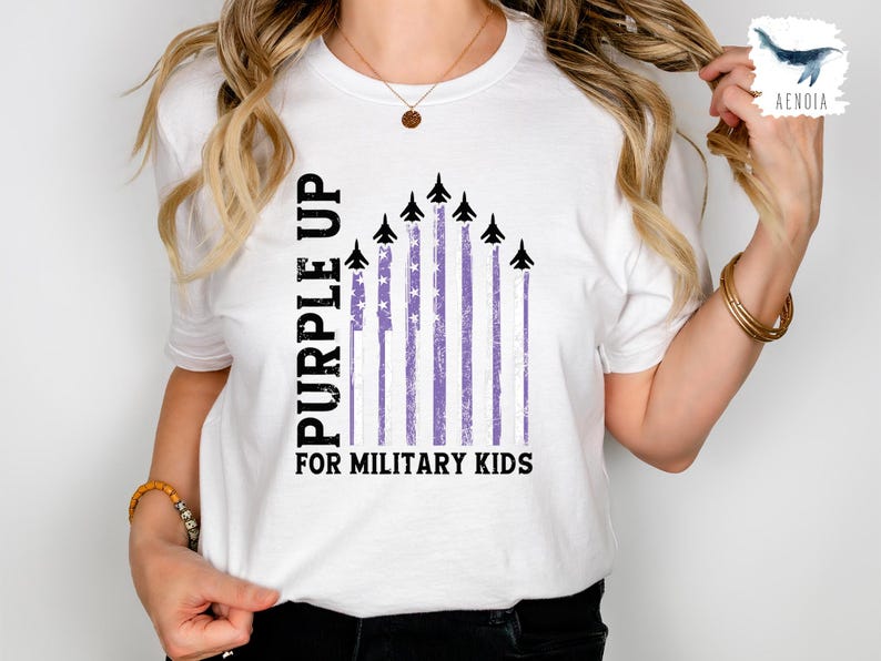 Op de afbeelding: Wit T-shirt met een paarse opdruk voor militaire kinderen. De opdruk toont een paarse Amerikaanse vlag met vliegtuigen die eroverheen vliegen.