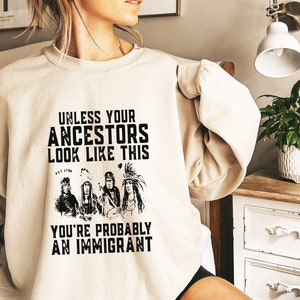 May include: A beige sweatshirt with a black graphic print of Native American people wearing traditional headdresses. The text on the sweatshirt reads "Unless your ancestors look like this you're probably an immigrant."