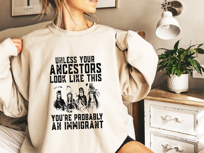 May include: A beige sweatshirt with a black graphic print of Native American people wearing traditional headdresses. The text on the sweatshirt reads "Unless your ancestors look like this you're probably an immigrant."