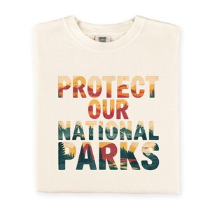 May include: A cream-coloured t-shirt with the words "PROTECT OUR NATIONAL PARKS" in a layered design. The text features a landscape of mountains, trees, and a sunset. The t-shirt is folded neatly.